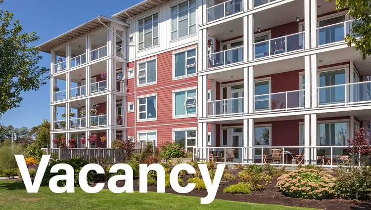 Modern red and white apartment building with balconies, landscaped garden, and large Vacancy sign in front